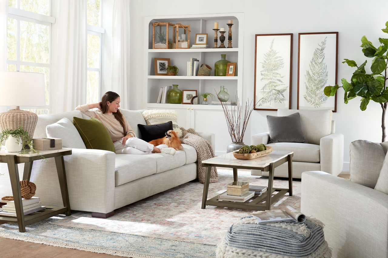 Destinations 3-Seat Sofa and Swivel Chairs in Convert Ivory and Emory Coffee Table and End Table in Weathered Sand and a model with a dog in a room scene.