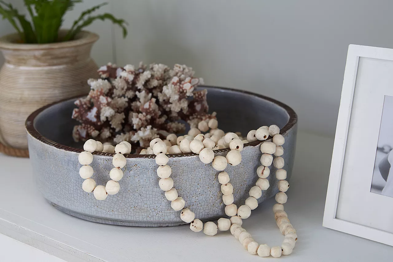 Closeup of a bowl with dried coral and a string of wood beads on a shelf in a room scene.