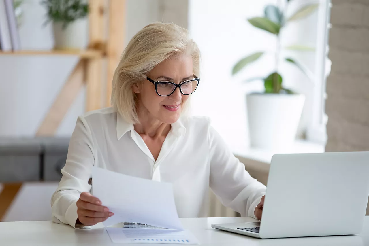 Aged businesswoman typing on pc holds paper prepare report analyzing work results feel satisfied do paperwork use business app online software for data analysis, financier manage firm finances concept