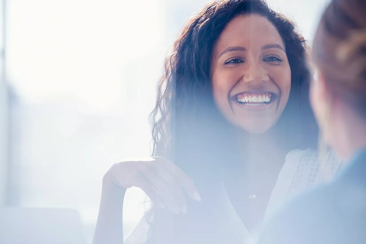 Business colleagues having a conversation. They are both young business people casually dressed in a modern office. Could be an interview or consultant working with a client. She is listening and smiling. One person has her back to us. Mixed ethnic group. One is African American and the other is Caucasian